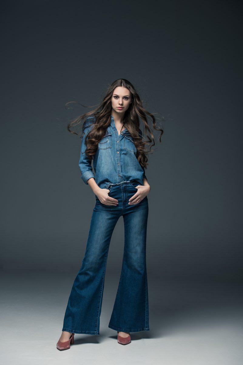 fashionable young woman girl with long hair posing in flare jeans, on grey