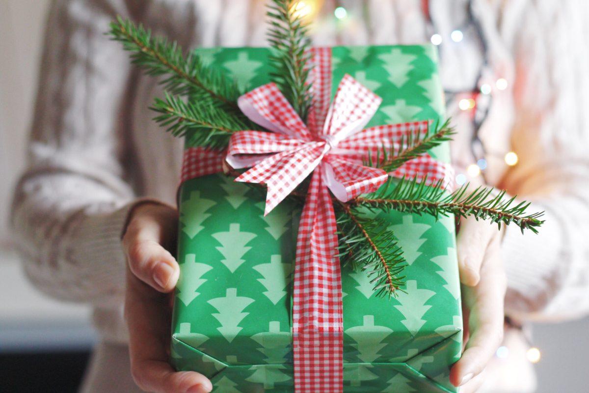 Person holding Christmas present wrapped in green paper with a pattern of green evergreen trees tied up with a red bow.