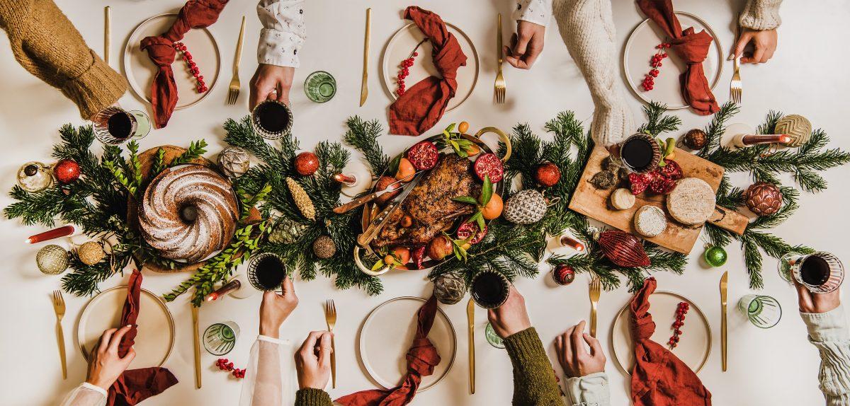 Flat-lay of Christmas table setting and people celebrating over dinnerware, cutlery, glassware, napkins, fir tree branches, roasted chicken, cheese board, bundt cake, candles over white background